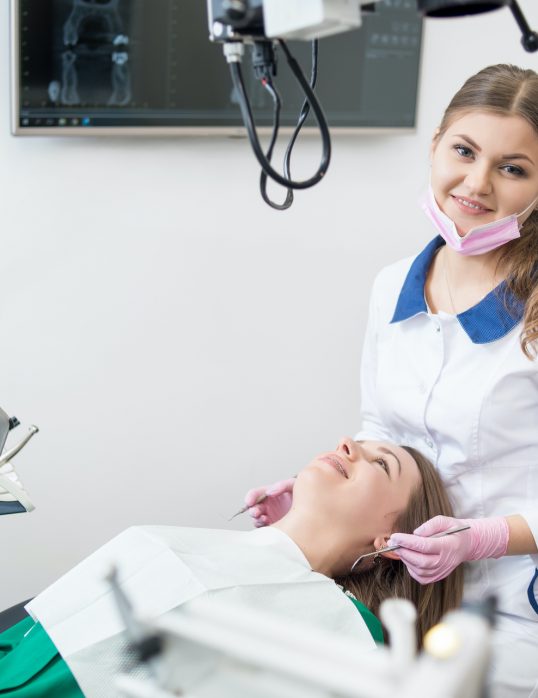 Friendly female dentist with happy patient during dental procedure in the dental clinic. Doctor looking to the camera, holding dental tools. Dentistry. Stomatology equipment