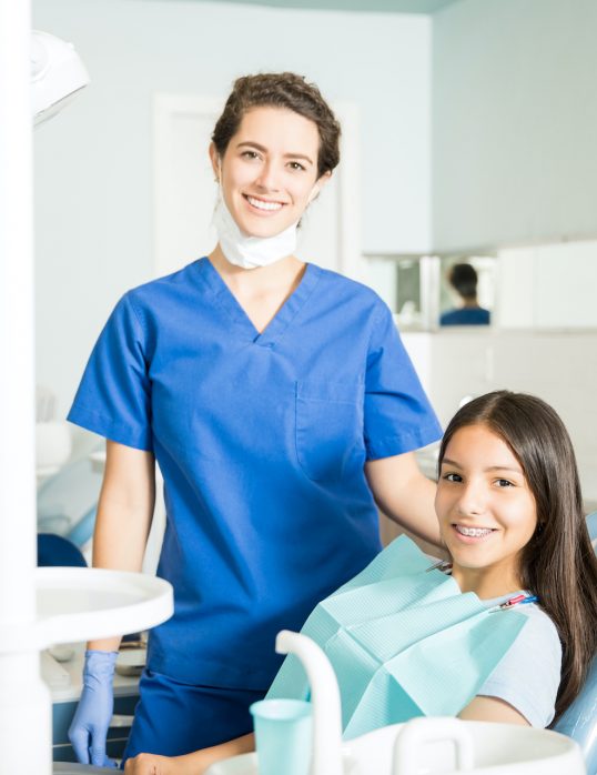 Portrait of smiling young female dentist standing by teenage girl in clinic